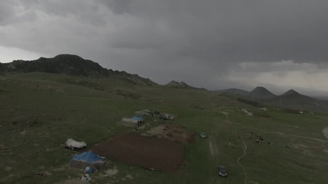 Dramatic aerial drone footage of a winding rural mountain road under a dark, stormy sky with heavy rain falling in the distance over the green valley hills landscape.

