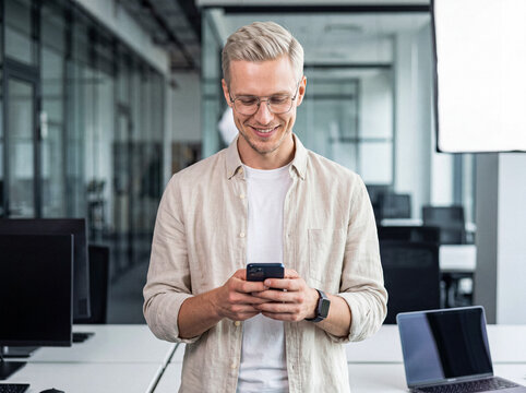 Smiling young businessman in glasses using a mobile phone in a bright modern office setting.
