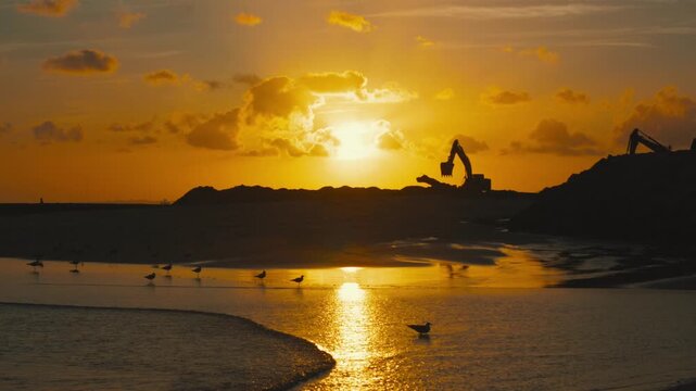 Sunset over beach with construction machinery silhouetted against the horizon, reflecting light on water and seagulls flying near the shore in a tranquil coastal scene