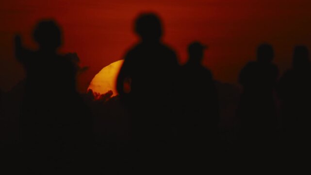 Silhouetted figures against a vibrant sunset, with the sun partially obscured by clouds, creating a dramatic scene of twilight in an outdoor setting