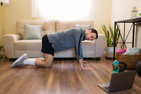 Hispanic man exercising at home following an online fitness class on a laptop, doing remote virtual workout in living room