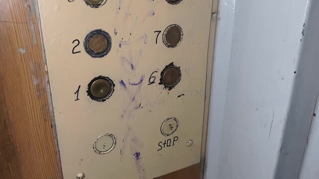 Panning close-up of a dirty vandalized elevator control panel with old worn buttons. Floor numbers are handwritten in black ink next to them to evoke a feeling of neglect.