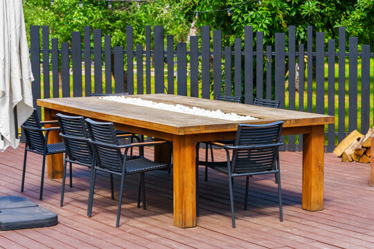 Large wooden outdoor dining table with black chairs on backyard patio terrace. Modern garden furniture for family gatherings, outdoor meals, entertaining guests and relaxing in landscaped yard.