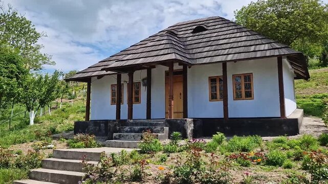 Traditional Moldovan peasant house with burned shingle roof. The cottage features white clay-plastered walls, wooden windows, and a porch supported by dark timber pillars, fronted by a blooming garden
