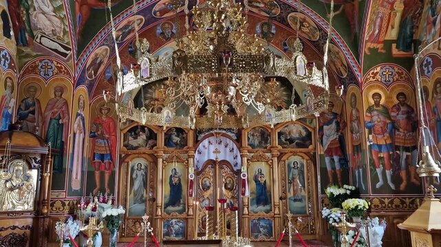 The inside view of Varzaresti Monastery with sacred icons resting on a white embroidered stand, revealing a grand golden iconostasis, a massive ornate chandelier, and breathtaking Biblical frescoes