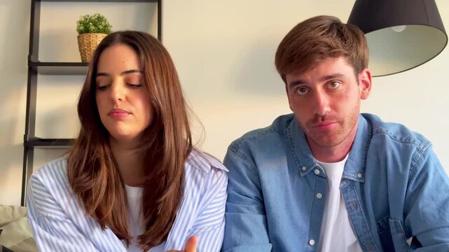 Millennial couple sitting on a sofa and having an intense discussion, showing a range of emotions from disagreement to frustration and misunderstanding in their living room at home