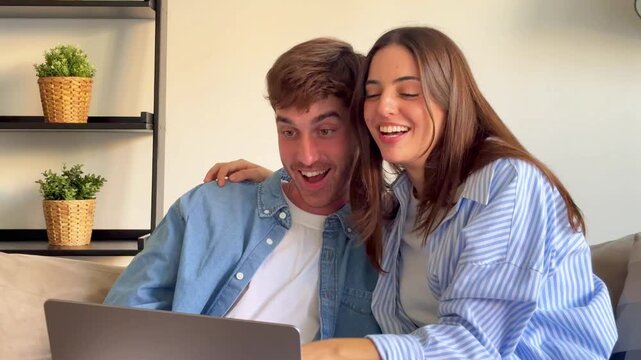Joyful couple sitting on the sofa at home while video calling family or friends on a laptop. Young man and woman waving and talking at the computer screen with happy expressions