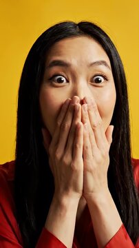 Surprised Asian Woman Covers Mouth with Hands on Yellow Background - A young Asian woman with wide eyes expresses shock or surprise by covering her mouth with both hands.