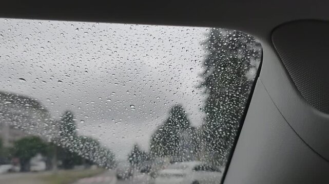 Raindrops accumulate on the glass windshield of a moving car while driving through a city street with blurry traffic ahead. This concept highlights dangerous driving conditions in rain.
