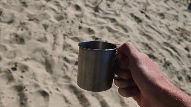 A male hand holds a stainless steel camping mug against a blurred background of bright sand texture. Concept of outdoor adventure, summer travel, and refreshing drinks in nature.