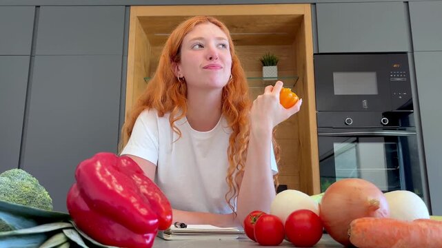 Joyful young redhead woman sitting at a kitchen table surrounded by fresh vegetables, smelling a yellow bell pepper and smiling while appreciating the aroma of healthy, organic food