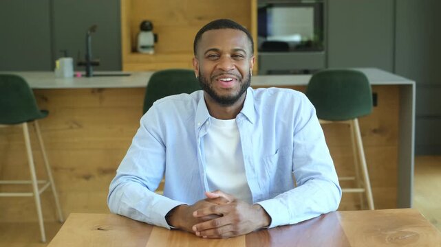 Young african american man smiling and talking while looking at the camera during a video conference, sitting at a wooden table at home with the kitchen in the background