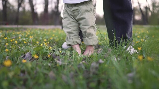Baby first steps on grass. Toddler learning to walk outdoors. Parent supports baby on grass. Exploring nature, baby steps on grass. Family moment in nature. Baby enjoying first walk in nature