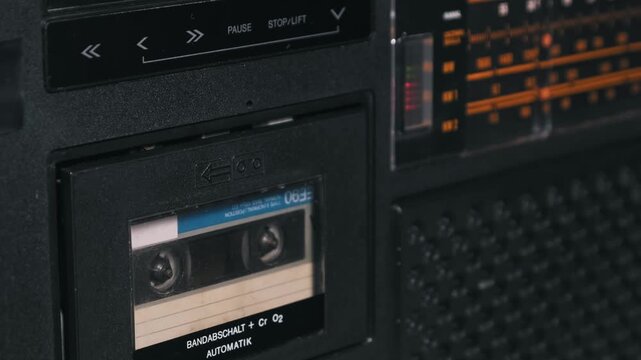 Close-up of a hand pressing the eject button on a retro black boombox to remove a cassette with a blue label. The closing of the empty tape deck highlights the internal mechanism of vintage tech.