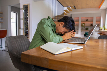 Obraz premium Asian man leaning over laptop, covering face with hands at dining table, notebook, green mug