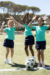 Diverse school-age girls jumping and raising arms on grass pitch with matching teal tees, balls © wavebreak3