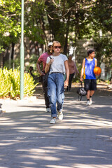 Fototapeta premium Diverse pedestrians walking down sunlit park path, woman wearing sunglasses carrying shoulder bag