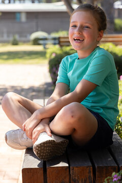 Female child sitting on bench at park, wearing teal shirt, holding white sneaker with knee scrape