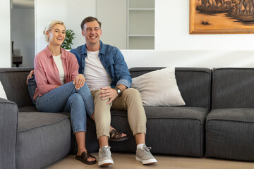 Obraz premium Couple sitting on dark gray sectional sofa in living room, man resting arm around woman smiling