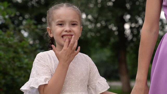 Playful little daughter shows gap in teeth holding mother hand in summer park. Funny daughter without lower teeth holds mother hand walking together in park. Family of mother and daughter in park