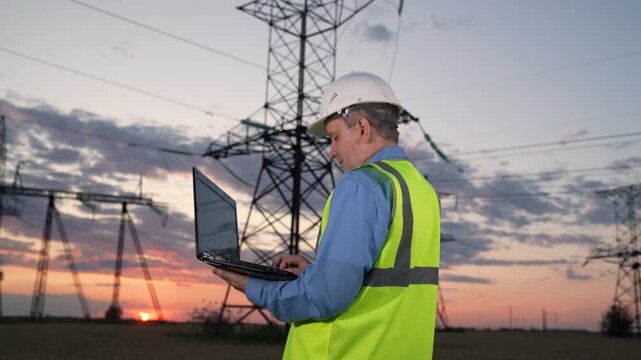 Man at power station, On-site engineering work, Monitoring electricity transmission, Working on laptop outside, Engineer and electrical towers, Industrial assessment, Technical field expert, Quality