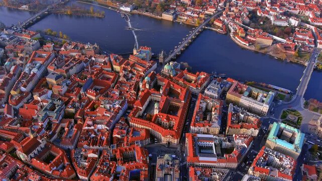 High angle drone footage captures Charles Bridge crossing the Vltava River in the historic Old Town of Prague, Czech Republic, featuring red tiled roofs during the golden hour sunset.