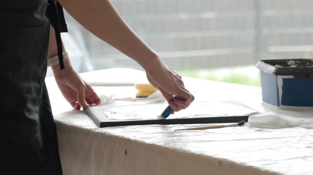 Woman creating textured artwork on canvas at home spreads white plaster paste with a palette knife at work table near a large window. Concept of DIY wall decor handmade art and creative hobby.