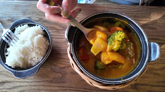 A person is using a spoon to serve a bowl of curry with vegetables, including broccoli and carrots, into a basket lined with a bamboo mat in Hoi An, Vietnam