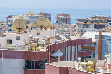 Fototapeta premium Rooftop solar water heaters with cylindrical tanks and panels and satellite dishes on a modern apartment building against a cityscape with a backdrop of the sea and clear blue sky