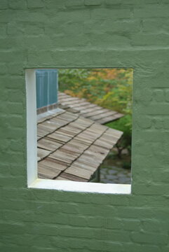 View of overlapping wooden shingle roofs through a square window on a green brick wall