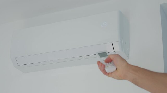 Hand adjusts air conditioner with remote control setting temperature near a white wall unit. Concept of cooling climate control and summer comfort.