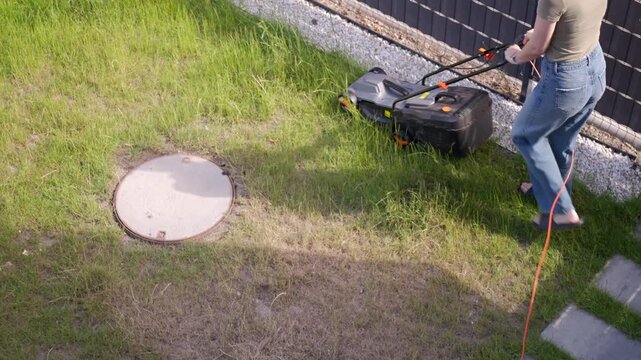 Woman mowing grass with an electric lawn mower in a backyard pushing the mower along a stone path with a red power cable on the ground