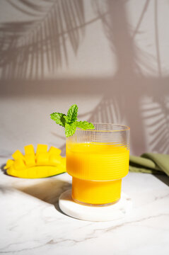 Glass of fresh mango smoothie and cut fruit on a white table with sun shadows.