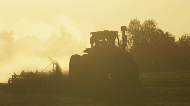 Tractor Silhouette Working In Dusty Field At Sunrise Or Sunset During Seeding, Soil Preparation And Farm Work In Rural Countryside