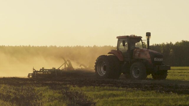Tractor Working Field In Dust Cloud At Sunset Agricultural Machinery During Evening Soil Preparation And Farming Operations