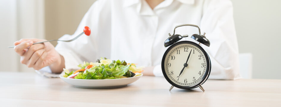 Intermittent fasting concept, close up clock, asain young woman, girl diet eating ketogenic low carb, fresh green vegetable salad on plate. Eat food healthy first meal in morning on table at home.