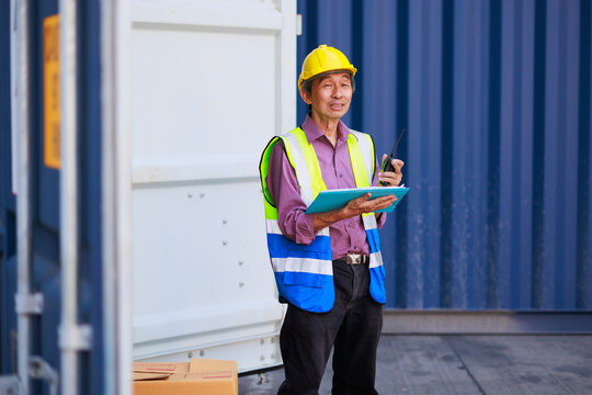 Asian senior foreman manager working through a radio communication with the workers in the container yard at port of import and export goods.