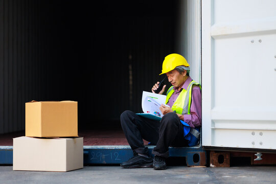 sitting on container box. Asian senior foreman manager working through a radio communication with the workers in the container yard at port of import and export goods.