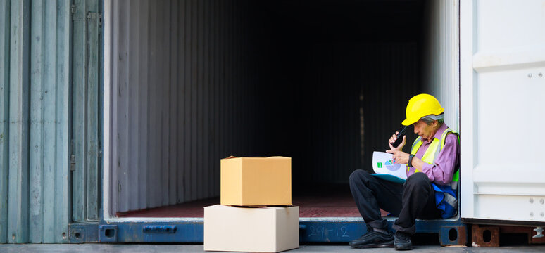 sitting on container box. Asian senior foreman manager working through a radio communication with the workers in the container yard at port of import and export goods.