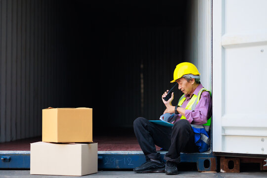 sitting on container box. Asian senior foreman manager working through a radio communication with the workers in the container yard at port of import and export goods.