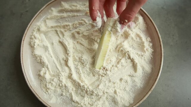 Close-up of fresh zucchini sticks being tossed and coated in plain white flour inside a shallow dish for crispy frying