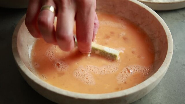 Close-up of fresh zucchini sticks being dipped and coated in whisked beaten egg inside a shallow dish for breading