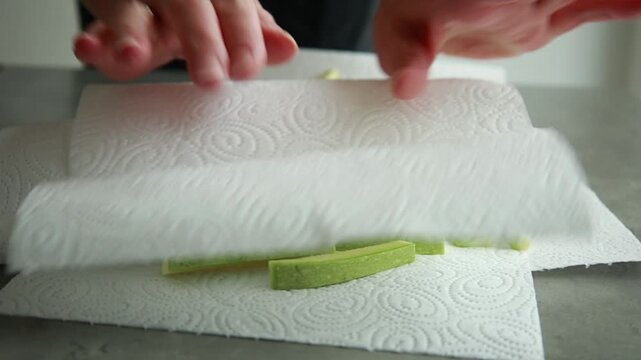 Close-up of a woman's hands gently patting raw zucchini fries with a white paper kitchen towel to remove excess moisture before breading