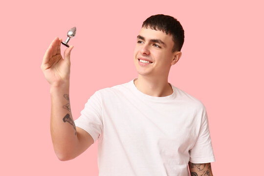 Smiling young man holding a small metal object with a shiny tip, looking upward on a pink background, he wears a white t-shirt and has tattoos on his arm