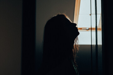 Silhouette of a young woman standing in a dim interior and facing a bright window. The image symbolizes hope, self-discovery, emotional healing and the search for clarity during difficult times. © vita