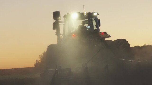 Farm Tractor Working Through Dust At Sunset With Sun Rays And Warm Golden Light Across Agricultural Field