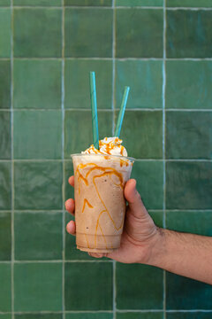 Hand holding a caramel coffee frappe with whipped cream against a green tile wall with copy space, summer iced drink