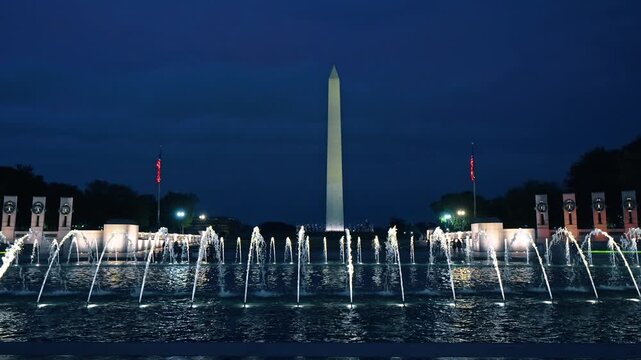 World War II Memorial fountains with Washington Monument at night, Washington, D.C. Illuminated water jets and state pillars framing distant obelisk under deep blue sky.