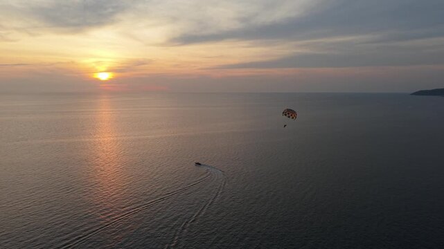 A parasailer is lifted into the air above the tranquil ocean during sunset, with a boat navigating below, creating a scenic view of the horizon and colorful sky