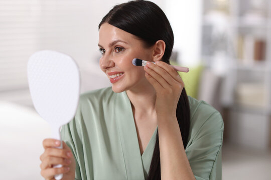 Young woman with mirror applying foundation with makeup brush indoors